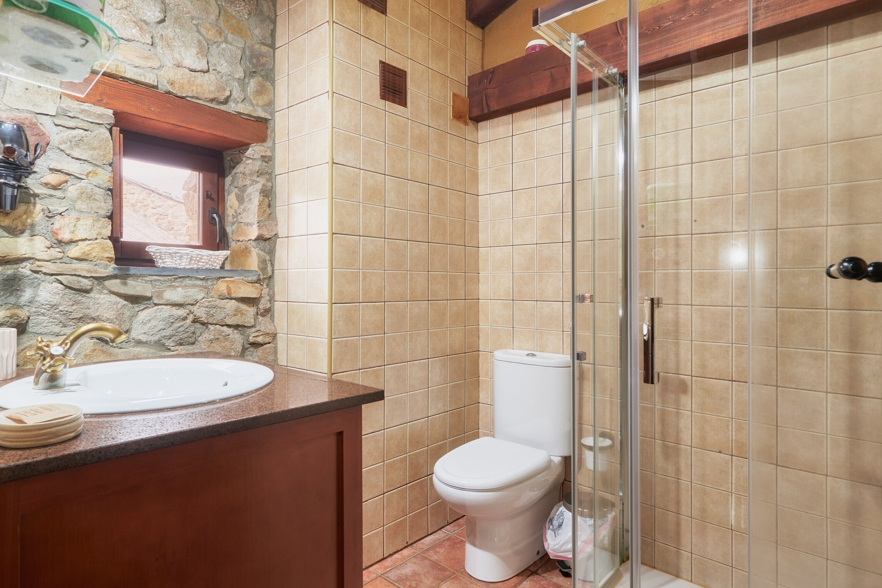 Un baño con pared de piedra, tocador de madera, lavabo blanco, inodoro y cabina de ducha de vidrio.