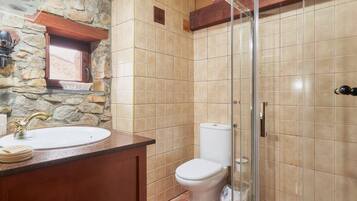 Un baño con pared de piedra, tocador de madera, lavabo blanco, inodoro y cabina de ducha de vidrio.
