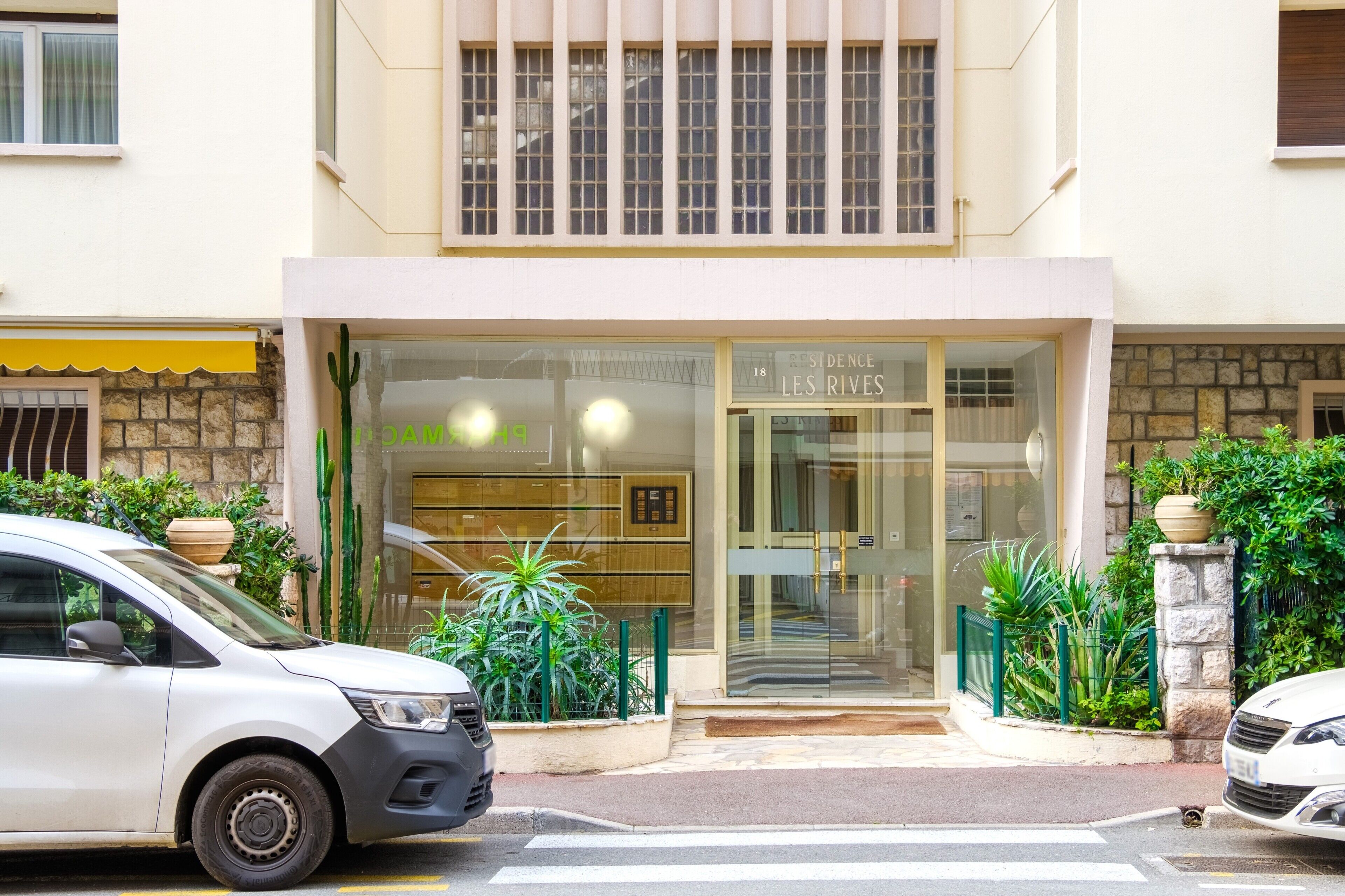 Entrada de hotel con fachada de vidrio, un coche estacionado frente a ella y una planta en maceta a la izquierda.