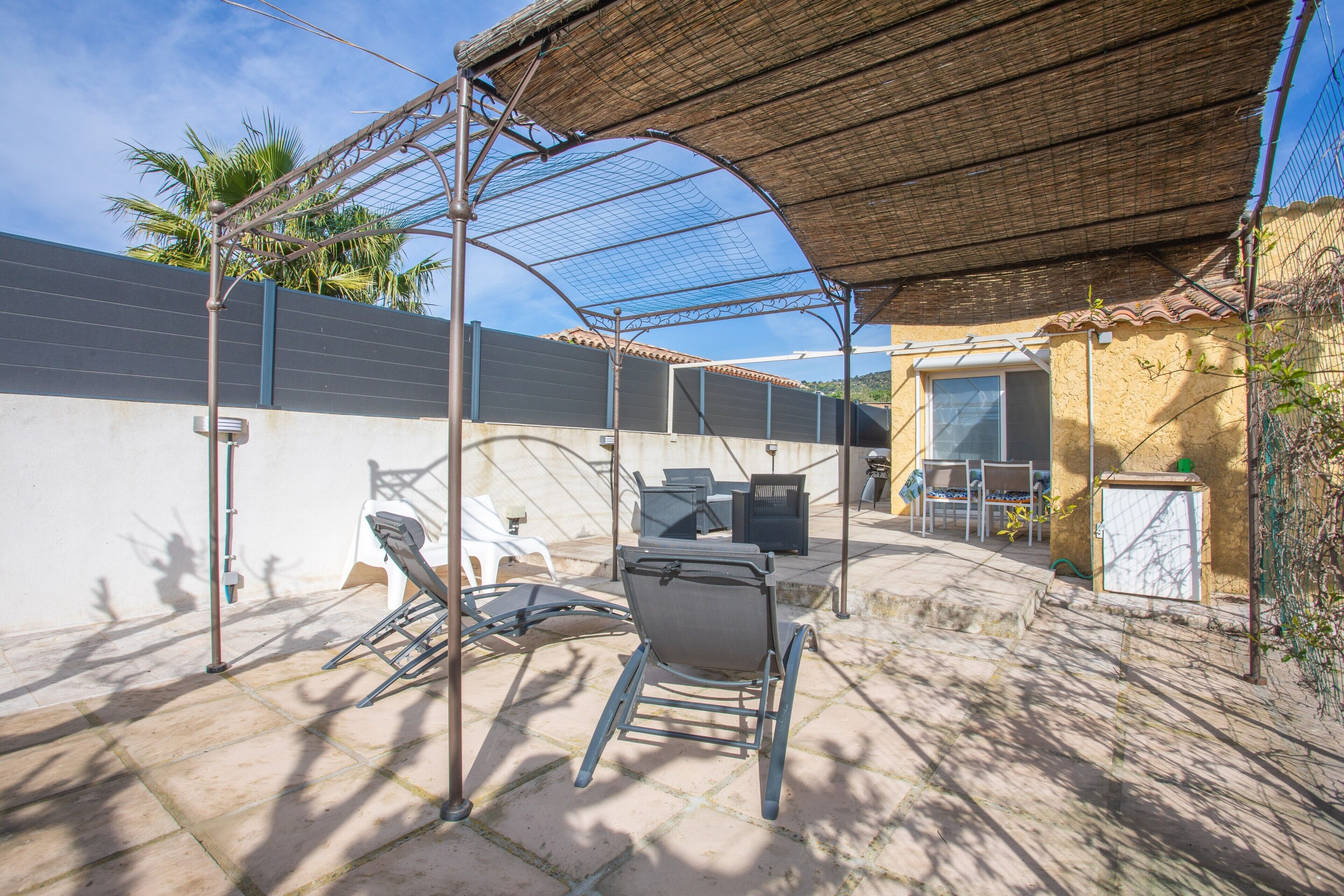 A covered outdoor seating area with chairs and a table, a building with a tiled roof, and a wall with a security camera.