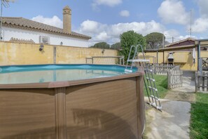 An outdoor swimming pool with a wooden deck, a ladder, and a white building with a tiled roof in the background.