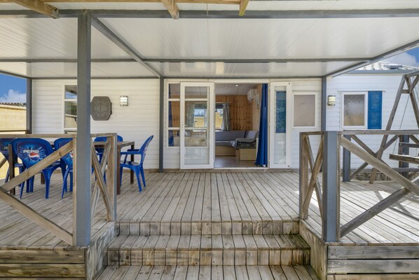 A covered outdoor area with a wooden deck, blue plastic chairs, and a visible room with a bed and curtains.
