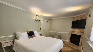 Une chambre à coucher avec un grand lit, une table de chevet, une chaise, une cheminée et un téléviseur fixé au mur.