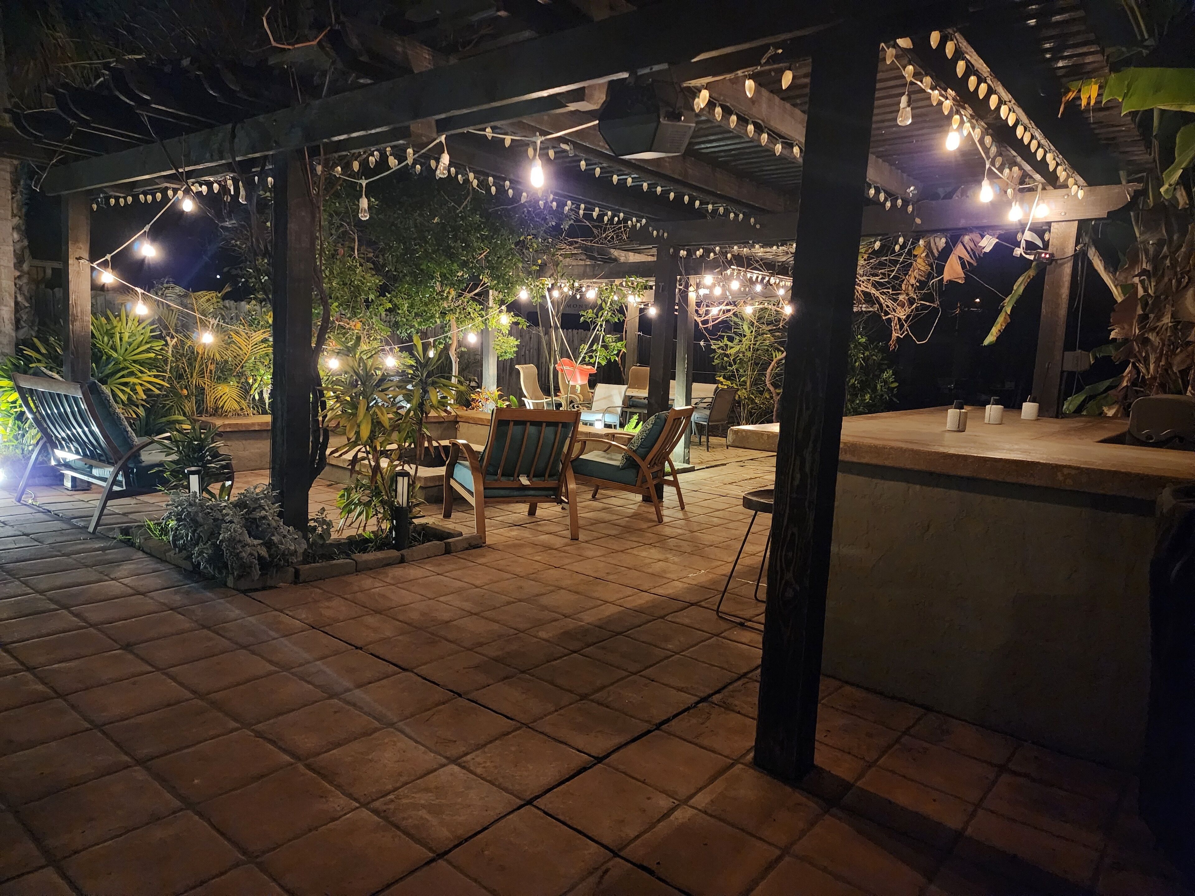 Un espace extérieur aménagé en terrasse, avec un sol carrelé, des chaises en bois et une pergola décorée de guirlandes lumineuses.