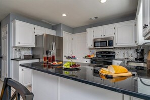 Una cocina moderna con armarios blancos, electrodomésticos de acero inoxidable y una isla central con frutas y pan.