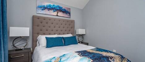 Een slaapkamer met een gestoffeerd hoofdeinde, een bed met witte beddengoed en blauwe kussens, een decoratieve plaid, nachtkastjes met lampen en een schilderij van een boom aan de muur.