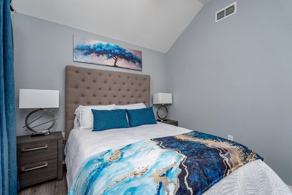 Un dormitorio con cabecera capitonada, cama con ropa de cama blanca y almohadas azules, manta decorativa, mesitas de noche con lámparas y un cuadro de un árbol en la pared.