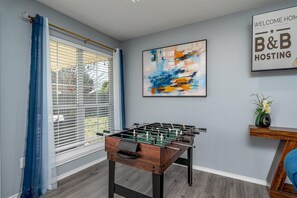 Une pièce avec une table de baby-foot, une fenêtre avec des stores, un tableau et une pancarte de bienvenue.