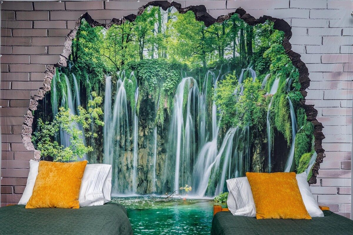 Un dormitorio con un gran mural en la pared de una cascada, dos camas con almohadas blancas y amarillas, y una pared de ladrillo.