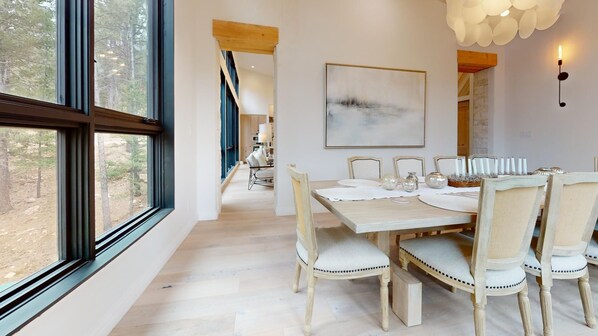 A dining area with a large table, chairs, and a view of the outdoors through large windows.