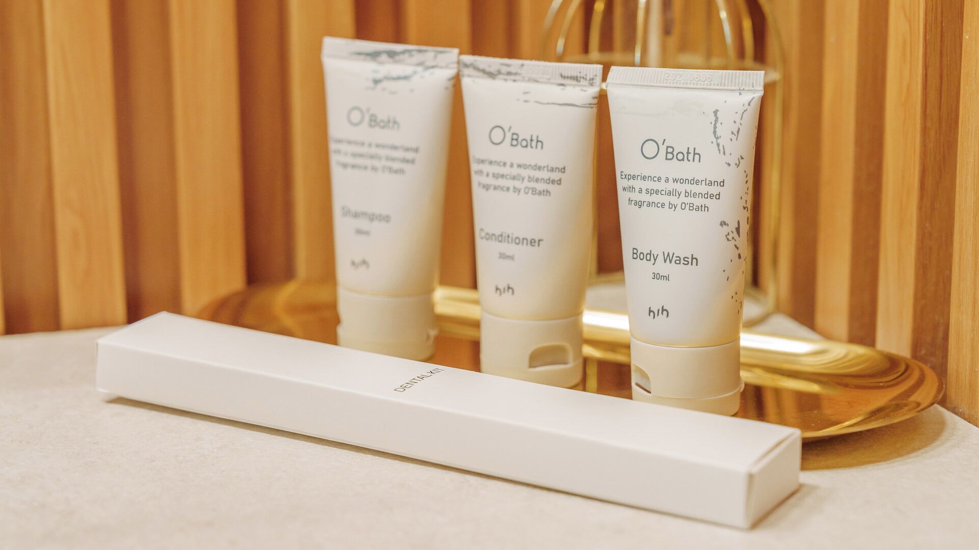 Tres productos de O'Bath: jabón corporal, acondicionador y champú, junto con una caja blanca, probablemente un kit de higiene bucal, sobre una superficie de madera.