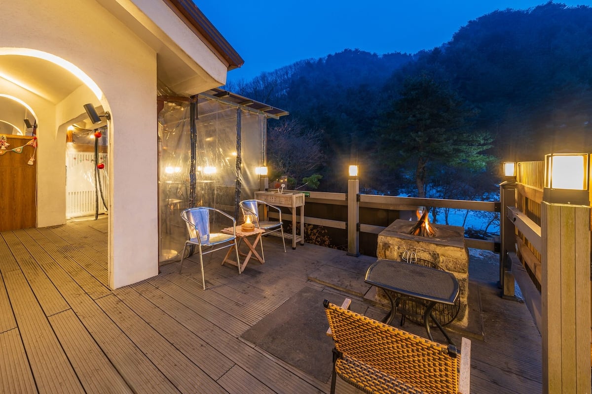 Una terraza de madera con mobiliario exterior, un hogar para fogata y vistas del bosque circundante.