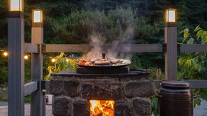 Un barbecue extérieur en pierre, avec un foyer, entouré d’une clôture en bois et illuminé de guirlandes lumineuses.