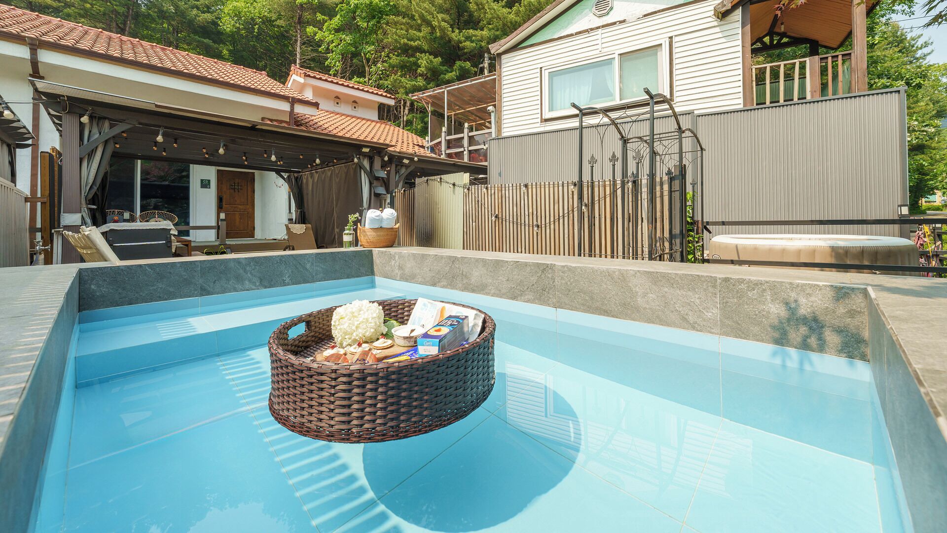 A swimming pool with a floating basket containing items, surrounded by a house with a covered patio and a fenced yard.