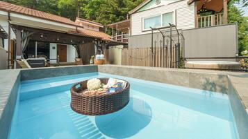 A swimming pool with a floating basket containing items, surrounded by a house with a covered patio and a fenced yard.