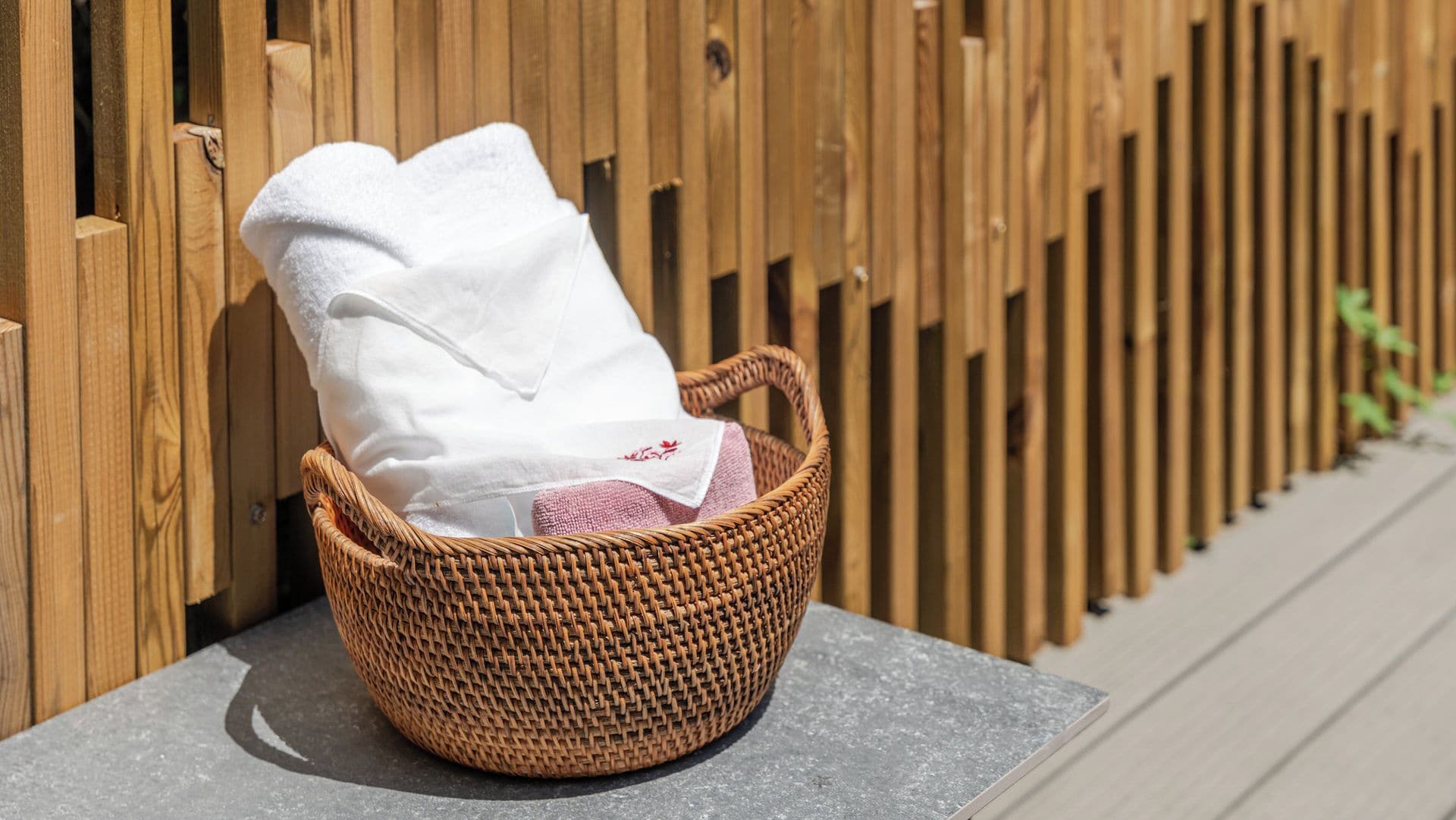 A wicker basket with a white towel and a pink cloth on a stone surface against a wooden fence.