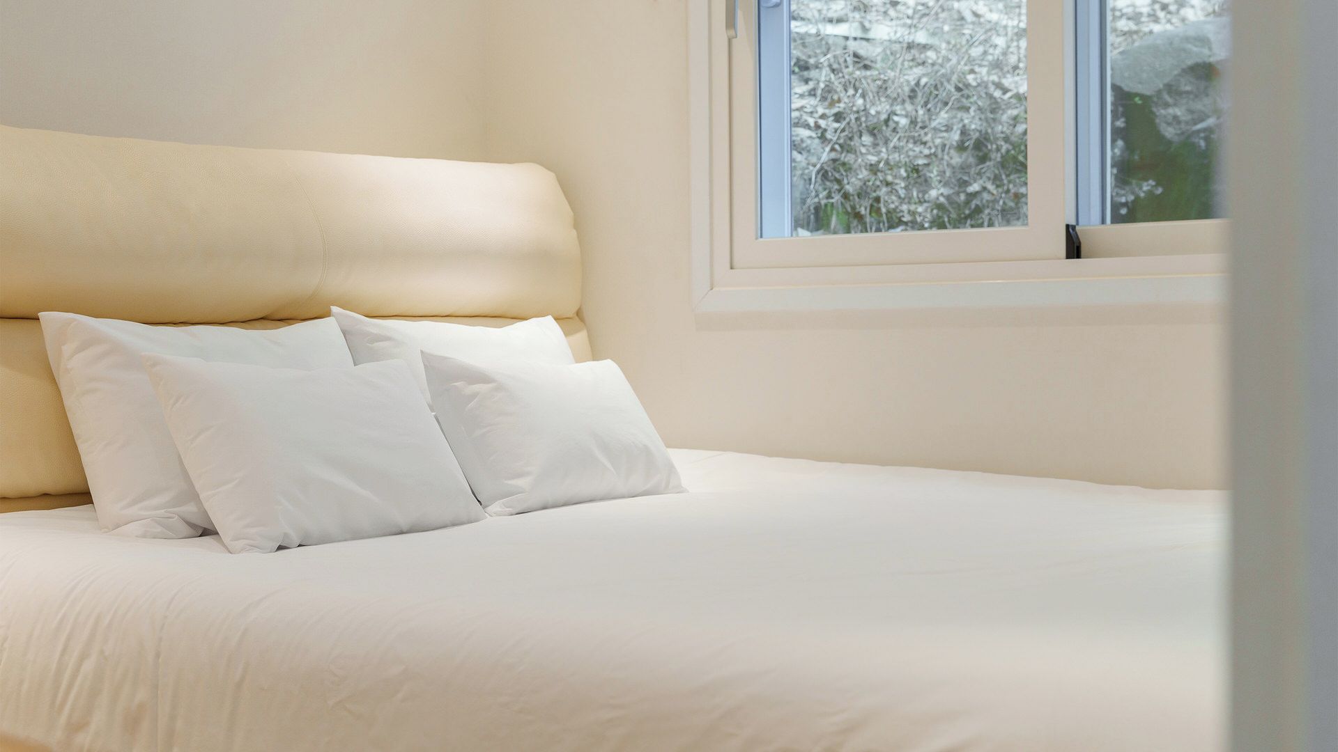 Una cama con ropa de cama y almohadas blancas, una ventana con vista a árboles y una pared sencilla.
