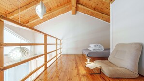 Une chambre à coucher aménagée dans un grenier, avec un plafond en pente, des poutres en bois, une fenêtre, un lit, une chaise et une lampe de lecture.