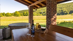 A wooden table with a bottle of wine, a glass, and a basket of snacks under a brick column with a view of a green field and hills. - Stunning home in Prvan Selo with sauna (Prvan Selo)
