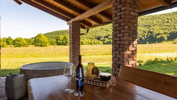 Een houten tafel met een fles wijn, een glas en een mandje met lekkernijen, onder een bakstenen pilaar, met uitzicht op een groen veld en heuvels.