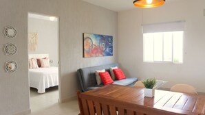 Una sala de estar moderna con una mesa de comedor de madera, un sofá gris con cojines rojos, un cuadro abstracto de colores y un amplio ventanal que proporciona luz natural.