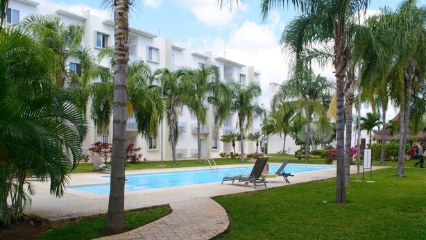 Zona de piscina con sillones de descanso, palmeras y un edificio con balcones al fondo.