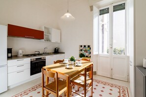 A compact kitchen with white cabinets, a red upper cabinet, a dining table with chairs, and a large window.