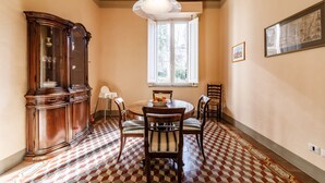 A dining room with a wooden cabinet, a round table with chairs, a window with a view, and a patterned floor. - GuestHost - Casa Calise - Elegant 1920s Villa w/ Garage (Siena)