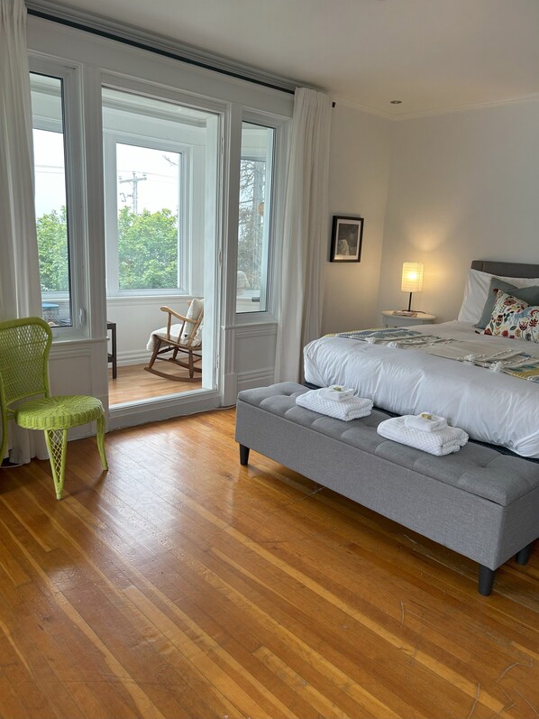 A bedroom with a grey sofa bed, a green wicker chair, a wooden rocking chair, and a view through a large window.