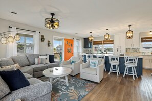 A modern living room with a grey sofa, white armchairs, and a kitchen area with white cabinets and blue bar stools.