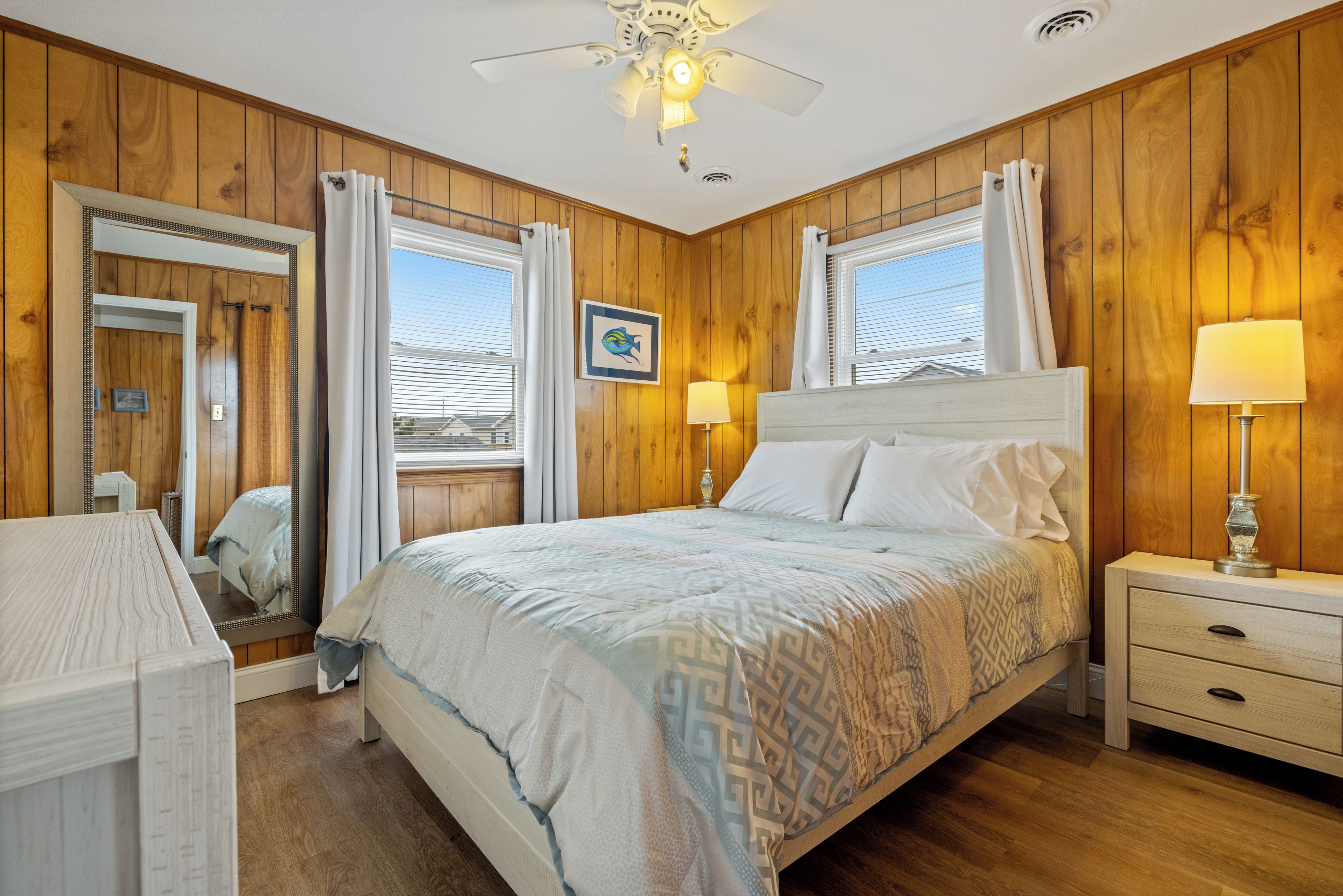 A bedroom with a wooden paneled wall, a bed with a patterned bedspread, a nightstand with a lamp, and a window with curtains.