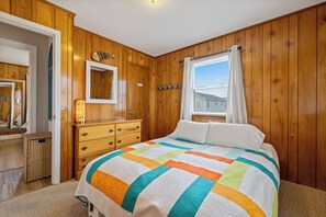 A bedroom with wooden paneling, a bed with a colorful quilt, a dresser, a mirror, and a window with curtains. - Happy Hour: Pet Friendly, Oceanside, Close to the Beach and Perfect for Families! (Kill Devil Hills)