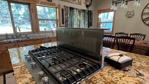 A kitchen with a granite countertop, a gas stove, wooden cabinets, and a window overlooking a forest.