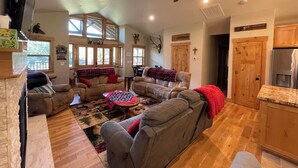 A spacious living room with a large sofa, a coffee table, and a fireplace. There is a ceiling fan, a kitchen area with wooden cabinets, and a dining table with chairs.