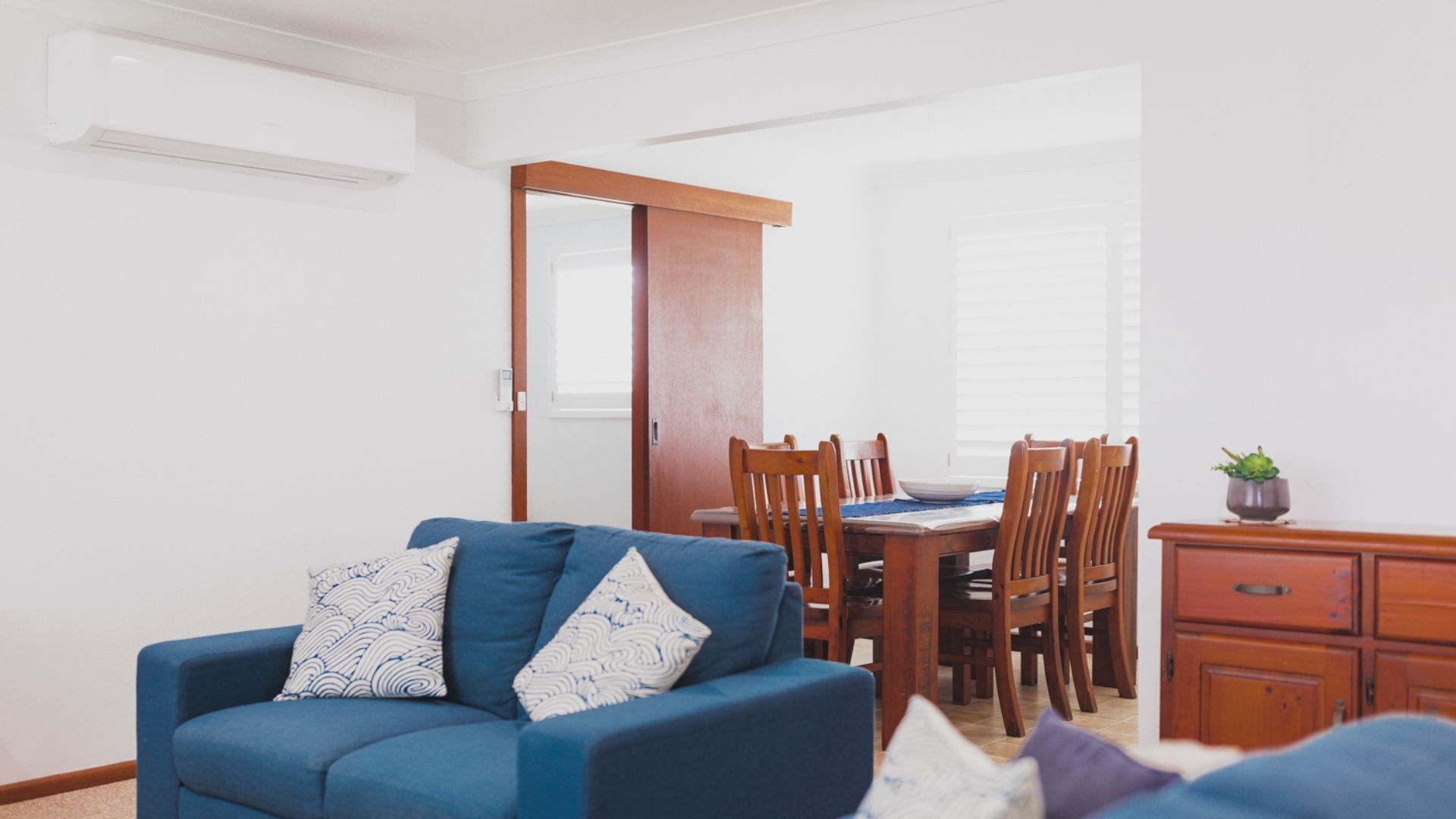 Una sala de estar con un sofá azul, una mesa de comedor de madera y sillas. Se ve un aparador de madera y una planta en maceta a la derecha.