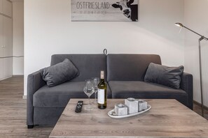 A modern living room with a grey sofa, a wooden coffee table, a remote control, wine glasses, and a bottle of wine. - 50165 Apartment Seestern - Small, but totally romantic: On approx. 45 sqm (Carolinensiel)