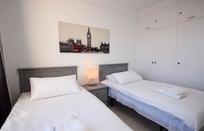 Une chambre d’hôtel avec deux lits simples, une table de chevet avec une lampe, un tableau représentant des monuments de Londres et une armoire avec des étiquettes indiquant « PRIVÉ ».