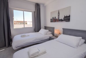 A hotel room with two beds, a window with curtains, a nightstand with a lamp, and a painting of a cityscape.
