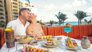 Ein Paar speist an einem Tisch mit Sushi und einem Cocktail auf einem Balkon mit Blick auf einen Strand.