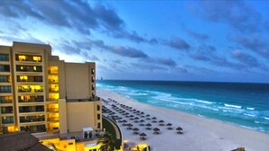 Ein Strandhotel mit mehreren Etagen, ein Sandstrand mit Sonnenschirmen und Meereswellen.