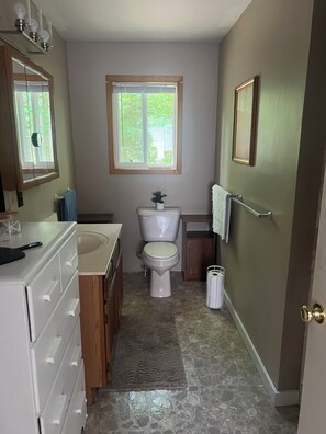 Un bagno con il water, un lavabo e uno specchio. C'è una finestra sopra il water e un portascopetti sulla parete.