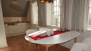 A dining area with a white table set for two, a kitchen in the background, and a window with curtains.