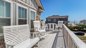 A wooden deck with white outdoor furniture, overlooking a beach and a house with a dark facade. - 4 br Ocean view, beach, pool! (Rodanthe)