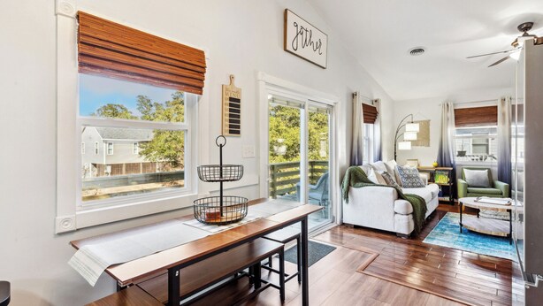 A spacious living area with a wooden dining table, a sofa, and a green armchair. There is a large window with a view of a house and trees.