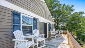 A wooden deck with white Adirondack chairs and a table, a house with beige siding and blue shutters, and a wooden fence.