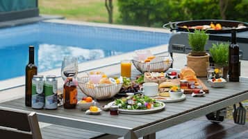 A poolside breakfast setup with various food items, drinks, and a grill.