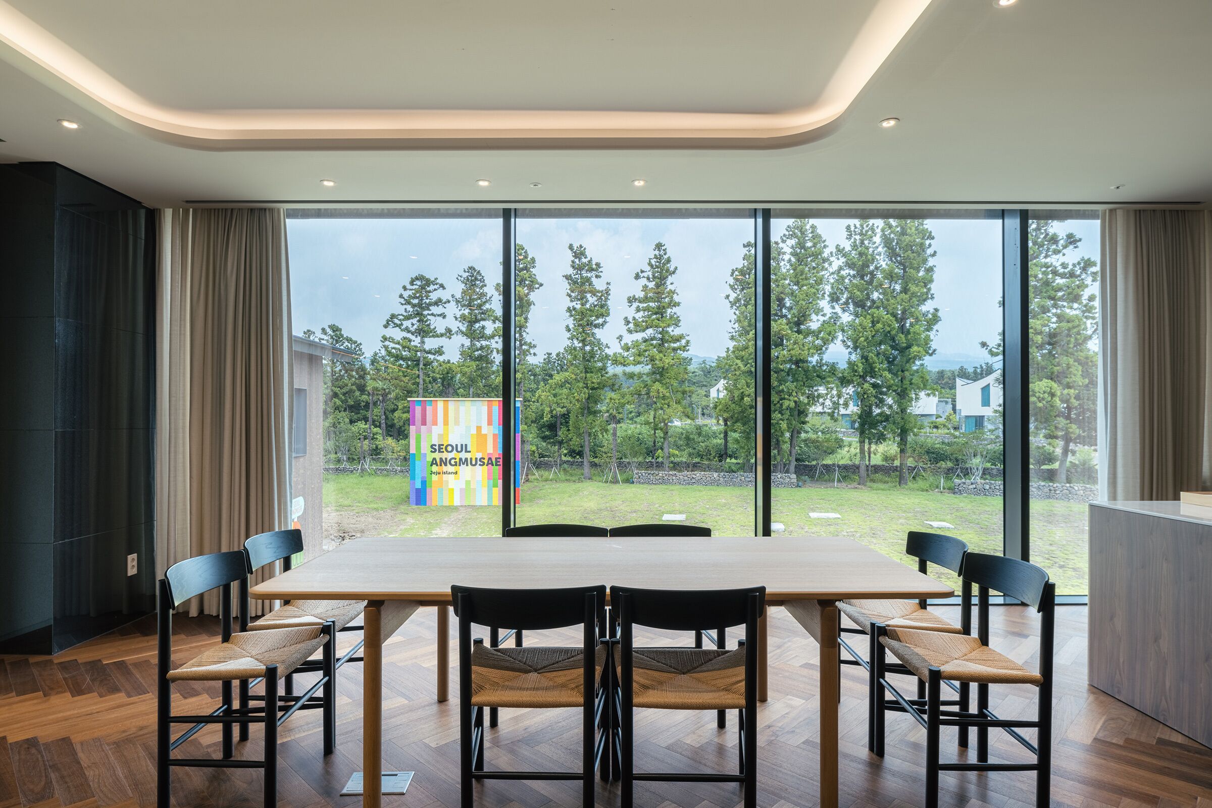 A modern dining area with a large table and chairs, large windows with a view of trees and a colorful sign.