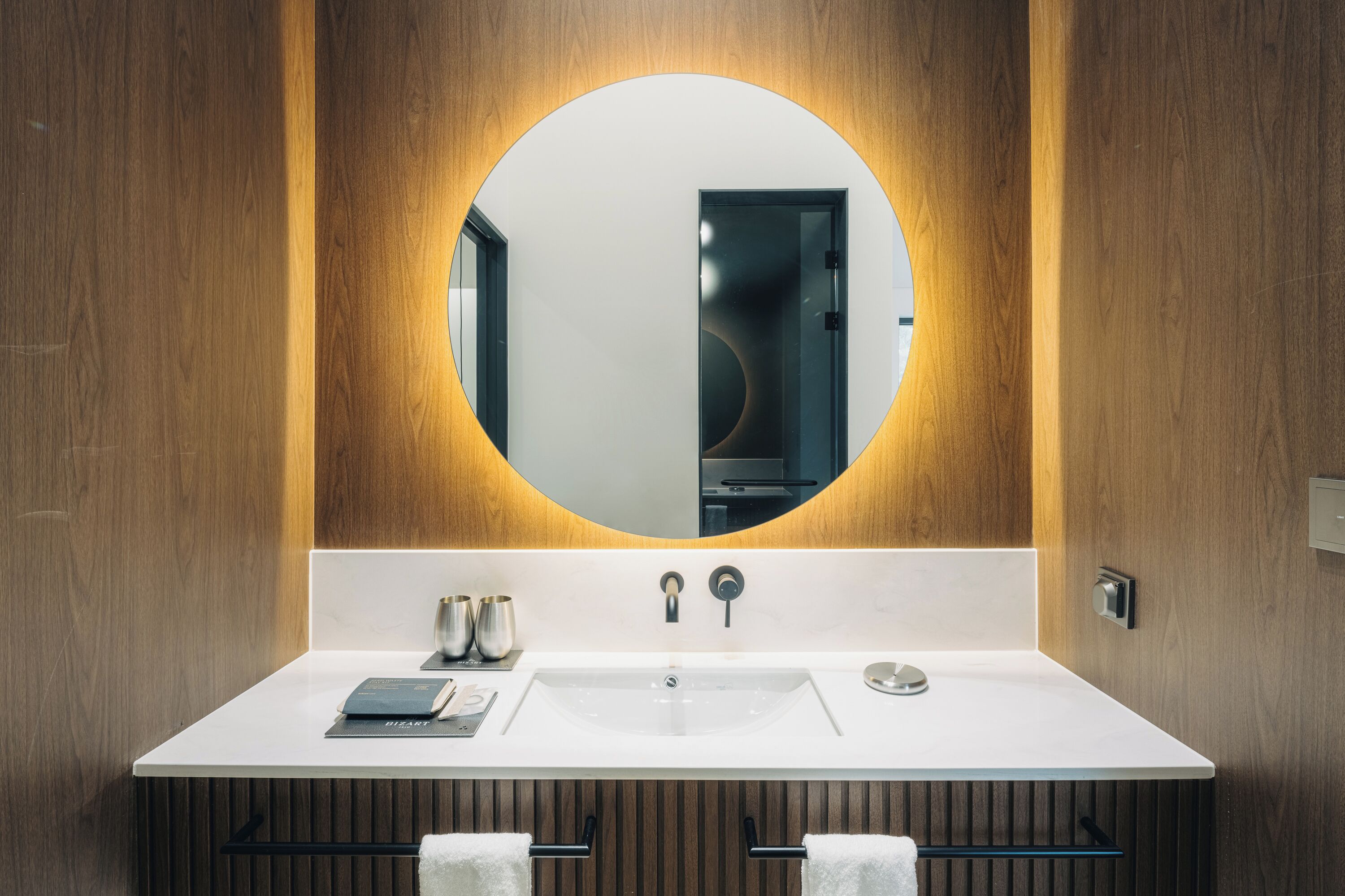 A modern bathroom with a large round mirror, a white sink, and wooden paneling.