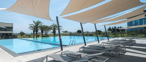 A swimming pool with lounge chairs and shade sails, surrounded by palm trees and modern buildings.