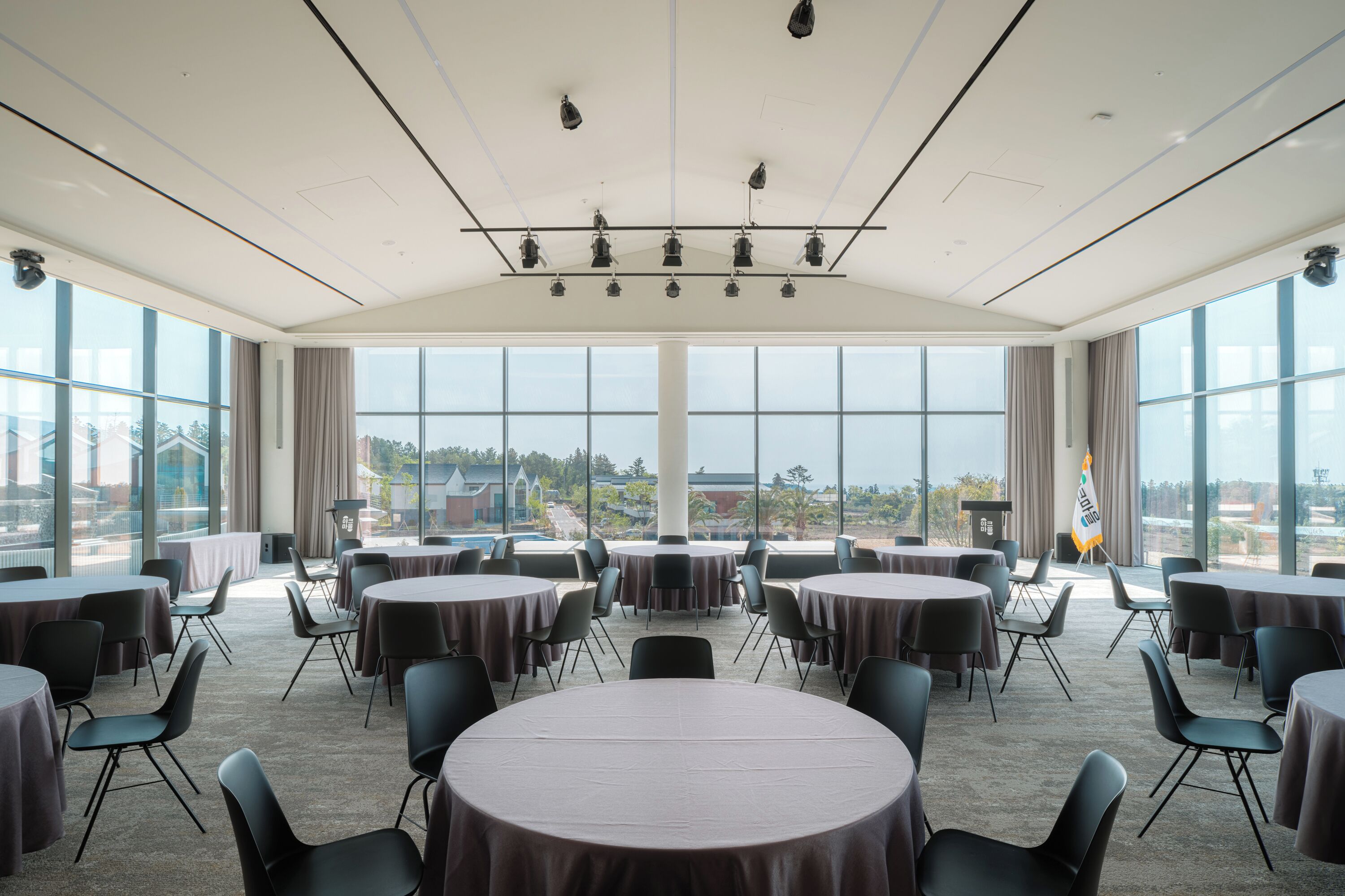A conference room with multiple tables and chairs, large windows offering a view of buildings and greenery, and a ceiling with lighting fixtures.